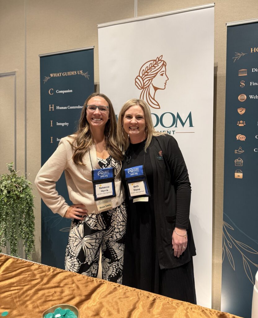 Rebecca Harris and Beth Diana smiling together in front of the Wisdom Management booth banners at the SDA conference.